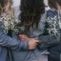 Giornata Internazionale della donna: in primo piano tra donne di schiena che si abbracciano cinte da fiori bianchi.